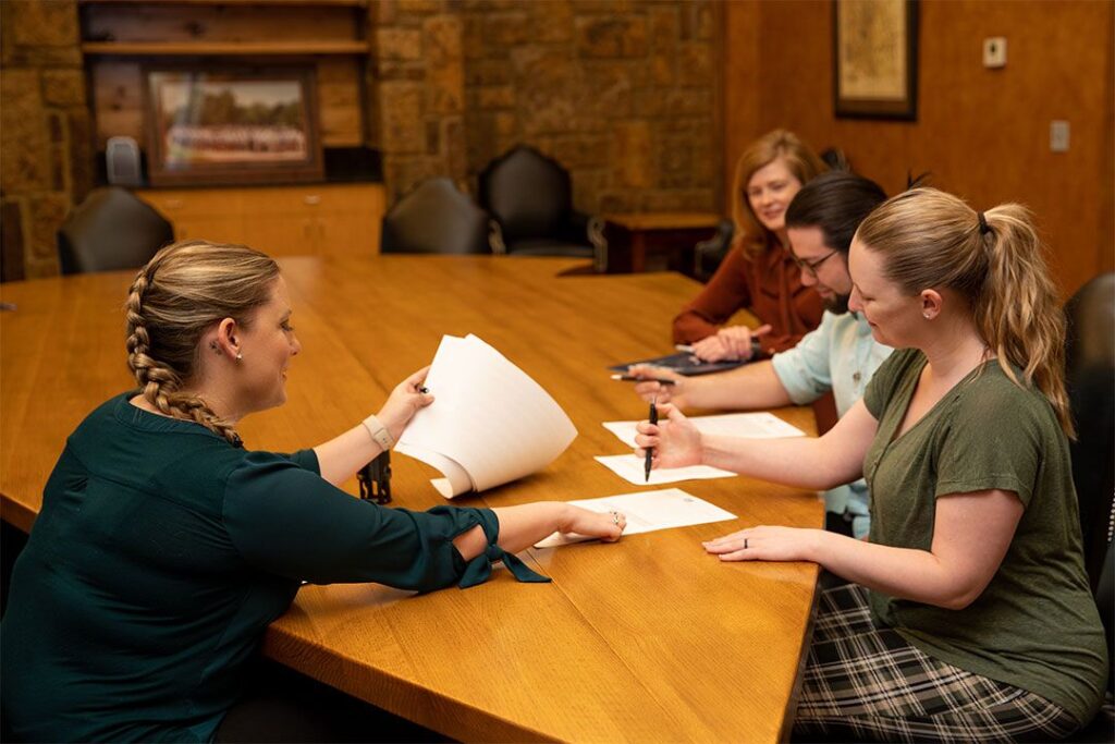 A group of 4 people sign papers at a table shaped like an arrowhead