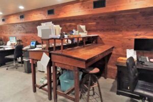 2 back to back wooden tables, as well as 2 other desks with computers and scattered paper on them